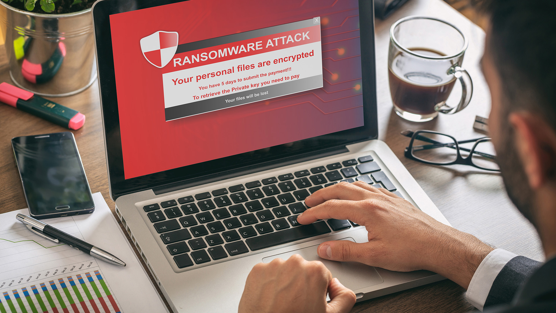 Laptop showing a red “Ransomware Attack—Your personal files are encrypted” warning while a person types—device lockout scenario.