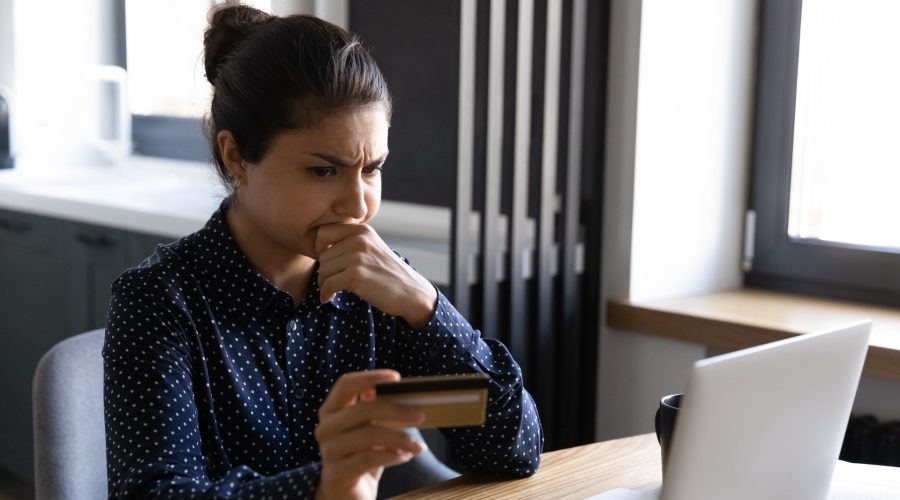 Woman sitting at a desk with a credit card and a laptop with a contemplative / unsure expression on her face. Spot and prevent check fraud
