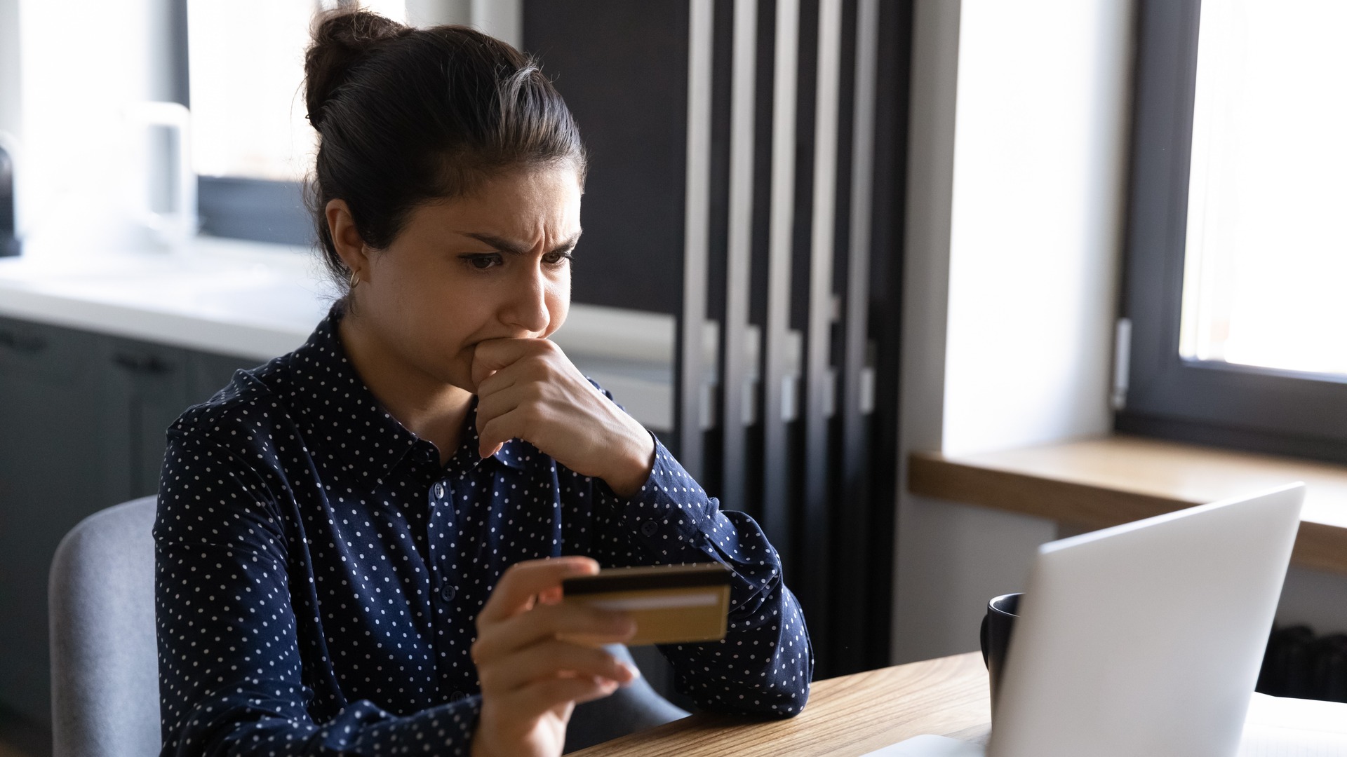 Woman sitting at a desk with a credit card and a laptop with a contemplative / unsure expression on her face. Spot and prevent check fraud